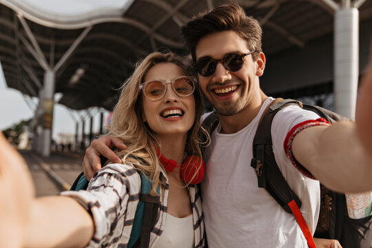 Couple if travelers takes selfie near airport. Young blonde curly woman in red headphones and brunette man in sunglasses smiles and hugs.