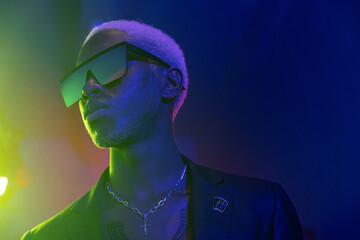 a guy with dark skin and blond hair. Dressed in a jacket and glasses that reflect green light. on his neck he had silver ornaments