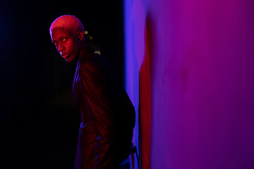 A guy with dark skin and white hair stands near the wall in a black suit. A purple light shines on his face - 445197008
