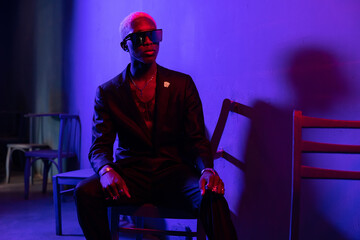 Obraz premium Dark-skinned guy sitting on a chair. Dressed in a black suit. on a background of purple light. He is wearing silver jewelry and black glasses