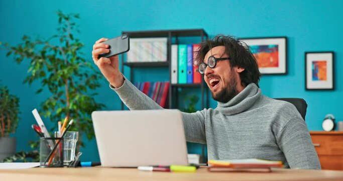 Funny boy with disheveled brown hair and big round black glasses sits at desk in office, taking selfie with phone, smiling broadly, showing thumb, bragging about new job among friends, on social media