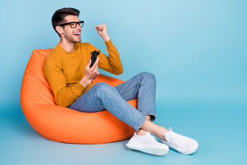 Portrait of attractive cheerful guy sitting in chair playing game having fun rejoicing isolated over bright blue color background