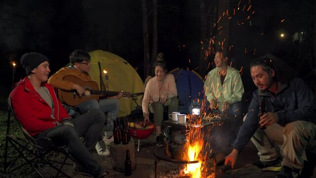 Group of Asian people sit on outdoor chair eating barbecue grill and beer for dinner with singing together at night. Man and woman friends enjoy outdoor lifestyle activity camping on summer vacation.