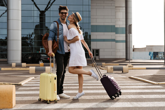 Attractive woman in white dress and hat kiss handsome brunette man. Cool tourists poses with suitcases near airport.