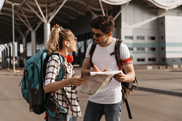 Blonde woman and brunette man looks at map and decide where to go. Portrait of travelers with backpacks near airport.