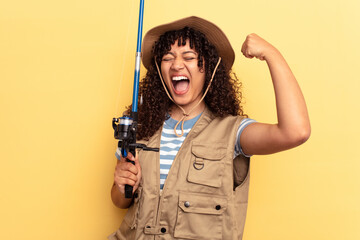 Young mixed race fisherwoman holding a rod isolated on yellow background raising fist after a...