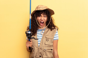 Young mixed race fisherwoman holding a rod isolated on yellow background screaming very angry and...