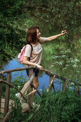 A happy young woman with short hair and a pink backpack stands on a small wooden bridge near the river in the forest, smiling and pointing into the distance. Selective Focus