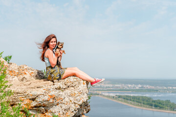 A young woman with a Yorkshire terrier sits on a cliff in the mountains in summer in front of a picturesque landscape with the Volga River