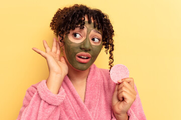 Young mixed race woman wearing a bathrobe holding a make-up remover sponge isolated on yellow background trying to listening a gossip.