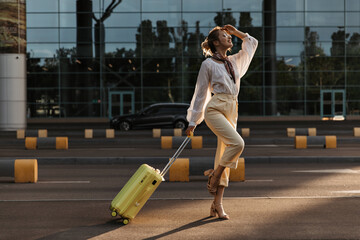 Charming young blonde woman in beige pants, white blouse and eyeglasses happily jumps near airport. Attractive lady in stylish outfit holds yellow suitcase.