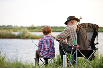 Rear view on senior man with grandson sitting near river with fishing rods in hands, enjoying beautiful nature, little boy learn to fish with grandfather. at weekends in evening, at sunset - Powered by Adobe