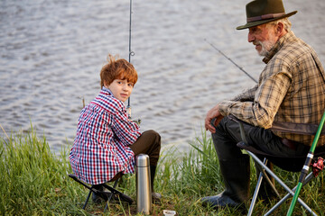 Mature caucasian man fishing with grandson, spending time near river, holding fishing rods in hands, enjoy spending time together, elderly man and little boy wearing checkered shirts. rear view