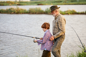 Side view on caucasian grandfather and grandson with fishing rods on river berth, friendly...