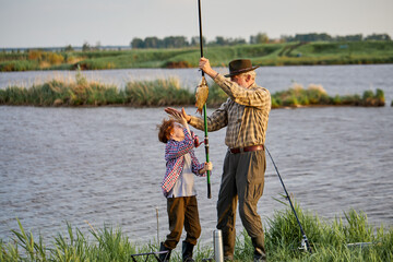 Summertime image of skilled fisherman on retirement having rest with grandson in wild nature using fishing rod, happy to catch fish, smile, give high five. Family, leisure and active hobby