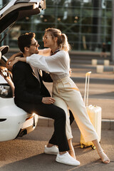 Blonde woman in white blouse, beige pants and eyeglasses hugs brunette boyfriend. Handsome guy in black suit sits in car and smiles.