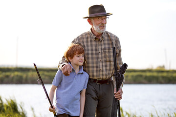 Portrait of senior man standing on the banks of lake with grandson holding fishing rod. family at weekends. handsome elderly man and kid boy standing near lake, engaged in fishing. copy space