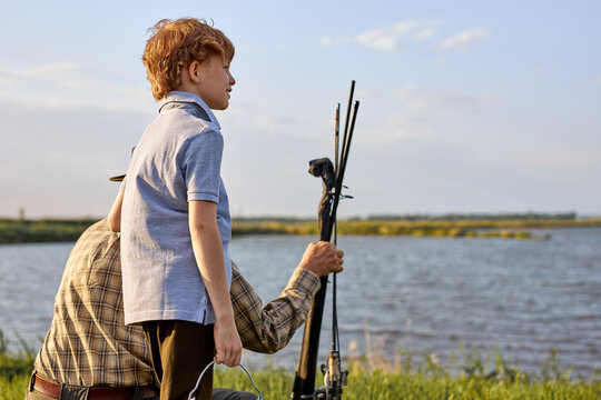 Adorable sweet redhead teenage boy came with granddad to fish, looking at side, looking forward to catch fish in lake, smiling. side view