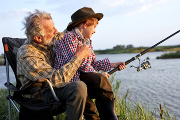 Mature man sitting on the banks of lake with grandson holding fishing rods. Little boy and adult man doing fishing as leisure activity, have talk, hobby while having spare time. in nature, countryside