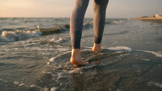 Slim female legs and feet walking along sea water waves on sandy beach. Pretty woman walks at seaside surf. Splashes of water and foam. Girl after running in fitness suit go on shore.