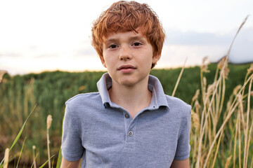 9 year old boy outside in grassy field, in countryside nature, look at camera, posing. Caucasian kid having natural red hair and freckles, in casual gray shirt. Portrait. Copy space. Children Concept © Roman