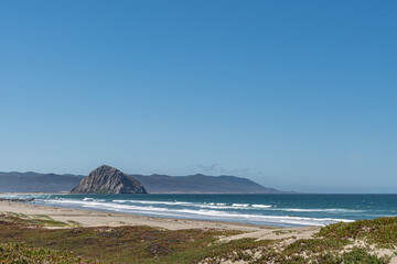 Morro Bay, CA, USA - June 10 2021: Dark gray Morro Rock in deep blue Pacific Ocean under blue sky with mountain range on horizon. Green covered dunes and sand in front. White surf. - Powered by Adobe