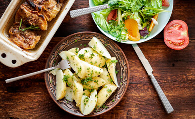 Boiled young potatoes with dill, fresh vegetable salad and baked chicken thighs on a dark wooden background.