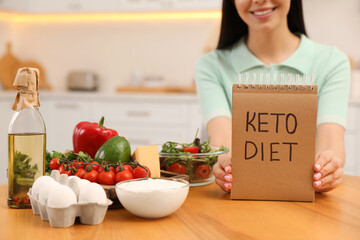 Happy woman holding notebook with words Keto Diet near different products in kitchen, closeup