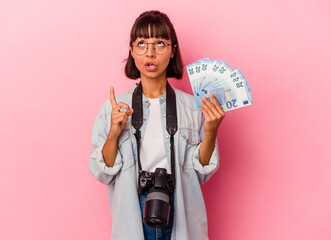 Young mixed race photographer woman holding bills isolated on pink background pointing upside with opened mouth.