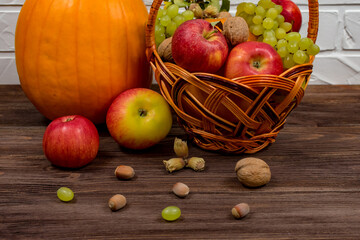 Orange pumpkins with apples and pears in a wicker basket with grapes, nuts and leaves on a brown wooden table against a white brick wall background. Close-up. Autumn still life.