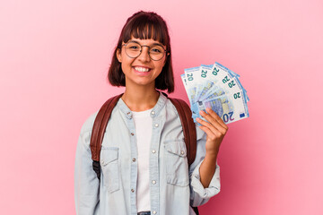 Young mixed race student woman holding bills isolated on pink background happy, smiling and...