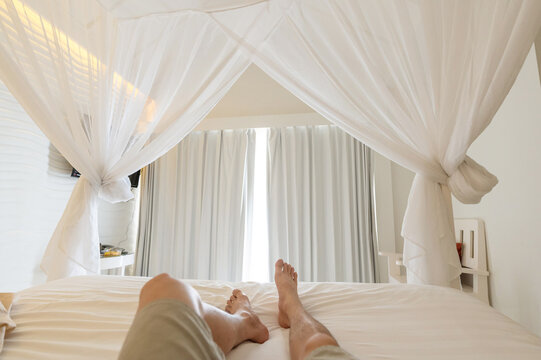 Man stretching legs in mosquito net on the bed at vacation, Sunlight through white curtain