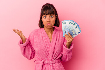 Young mixed race woman wearing a bathrobe holding bills isolated on pink background shrugs...
