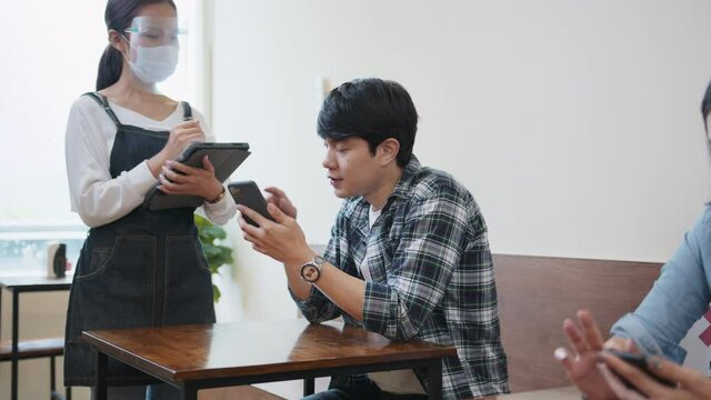 Young Asian man order food from restaurant waitress, use smartphone scan QR code for menu. Female customer sit in distance, social distancing sign. Coronavirus pandemic new normal lifestyle concept