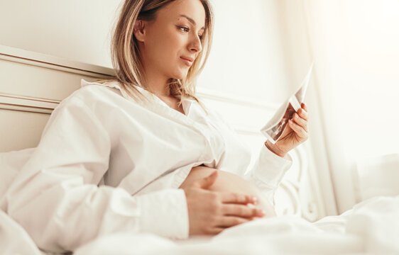 Happy pregnant woman with ultrasound scan resting in bedroom