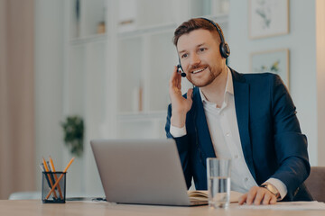 Break at work. Glad male entrepreneur wears headset on ears poses at desktop with laptop computer satisfied with his business achievements listens webinar online makes call via internet application