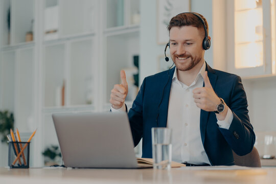 Happy male employee makes okay like gesture keeps thumbs up satisfied with colleague business presentation makes video call uses headphones with microphone wears formal clothing. Executive worker