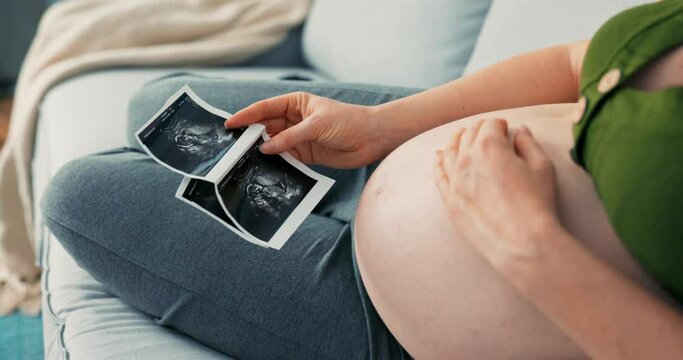 Shot from above with view of pregnant girl sitting comfortably on sofa, wearing sweatpants and short top, women touches large belly, puts hand on it, strokes it, looks at usg photo, ultrasound