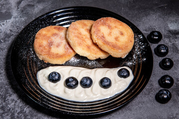 Cheesecakes (pancakes) with sour cream and blueberries on a black plate. Healthy tasty breakfast. Close-up
