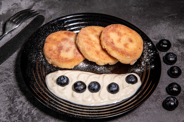 Cheesecakes (pancakes) with sour cream and blueberries on a black plate. Healthy tasty breakfast. Close-up
