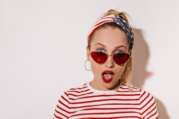 Fashionable girl with modern headband, gold earrings and red sunglasses in striped stylish t-shirt looking in surprise into camera on white backdrop..