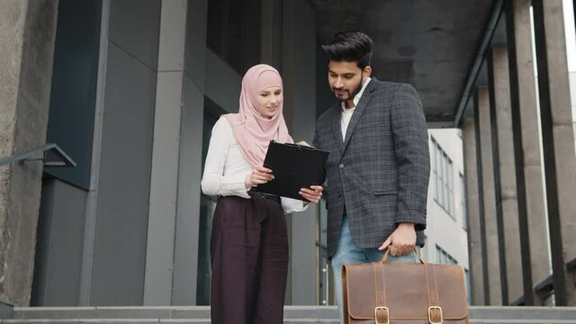 Beautiful woman in hijab holding clipboard and showing some documents to muslim man with suitcase. Two business partners standing outdoors and soling working issues. Arabian colleagues