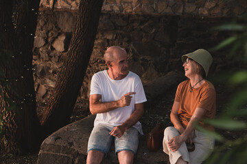 Happy senior couple (Asian woman and Caucasian man) sitting in tree shadow in city park and having a talk and laughing