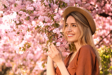 Young woman wearing stylish outfit near blossoming sakura in park. Fashionable spring look