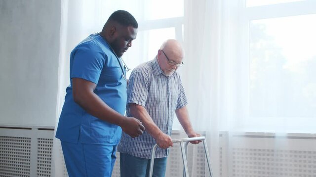 African-American caregiver is teaching disabled old man to walk with walker. Professional nurse and handicapped patient in a nursing home. Assistance, rehabilitation and health care.