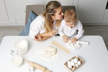 Happy mother and child in the white kitchen at the table put marshmallows on sticks, they eat it and they have a lot of fun