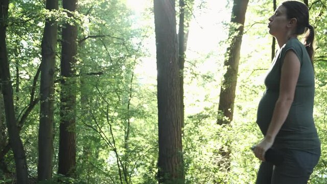 Active pregnant woman runs in summer park, stops to catch her breath. Expectant mother exercising healthy lifestyle jogging workout outside. Pregnancy sport and fitness training concept.