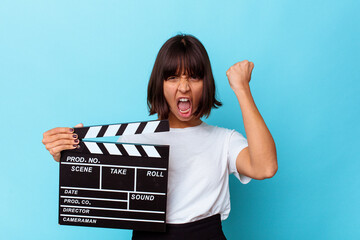 Young mixed race woman holding a clapper board raising fist after a victory, winner concept.