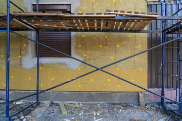 Fragment of a building surrounded by scaffolding. The wall is covered with sheets of green mineral wool for insulation. There is a window covered with brown roller shutters. Repair phase.