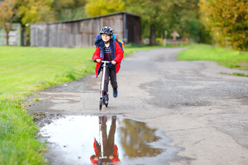 Fototapeta na wymiar cute little school kid boy riding on push scooter on the way to or from school. Schoolboy of 7 years driving through rain puddle. funny happy child in colorful fashion clothes and with helmet.